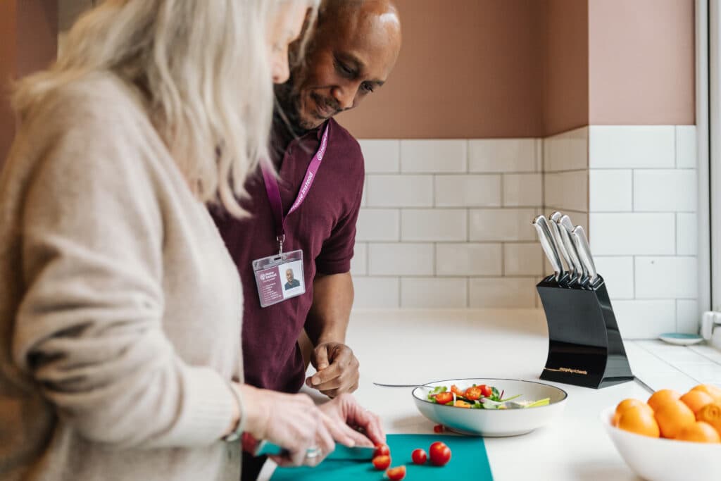 An older woman and a man prepare a salad together in a bright kitchen with a knife block nearby. - Home Instead