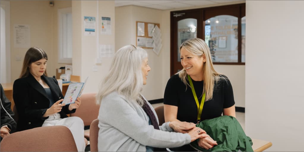 A woman with a lanyard smiles and holds hands with an older woman in a waiting room. - Home Instead