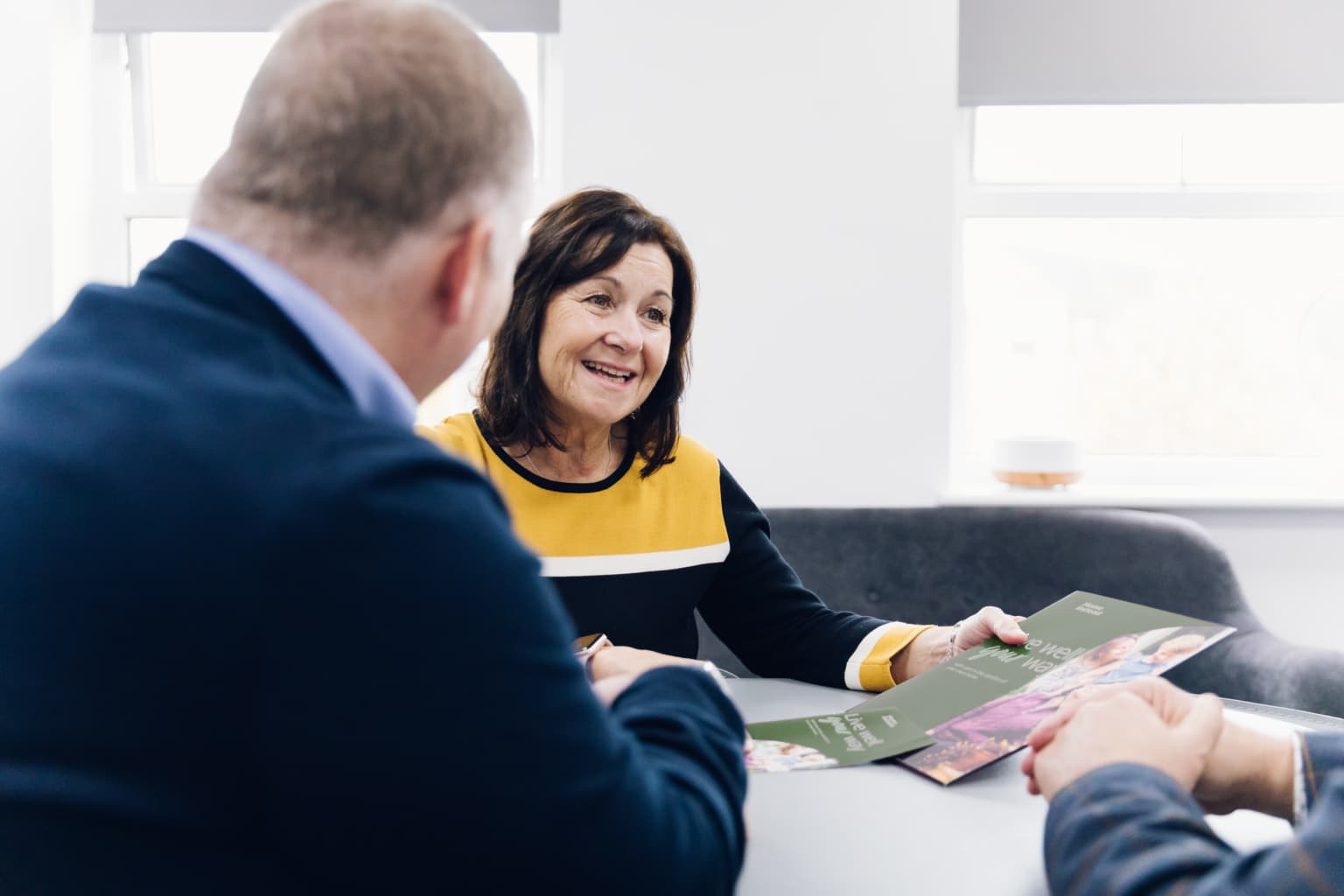 A woman smiling and holding leaflets while talking with two people at a table in a bright office. - Home Instead