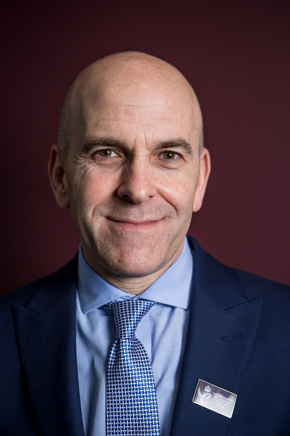 Bald man in a blue suit and tie smiling, standing in front of a dark red background. - Home Instead