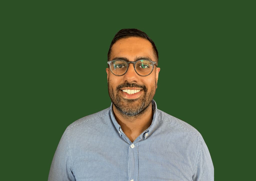 A man wearing glasses and a blue shirt smiles in front of a solid green background. - Home Instead