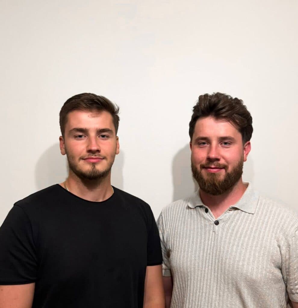 Two young men standing side by side and smiling in front of a plain white wall. - Home Instead