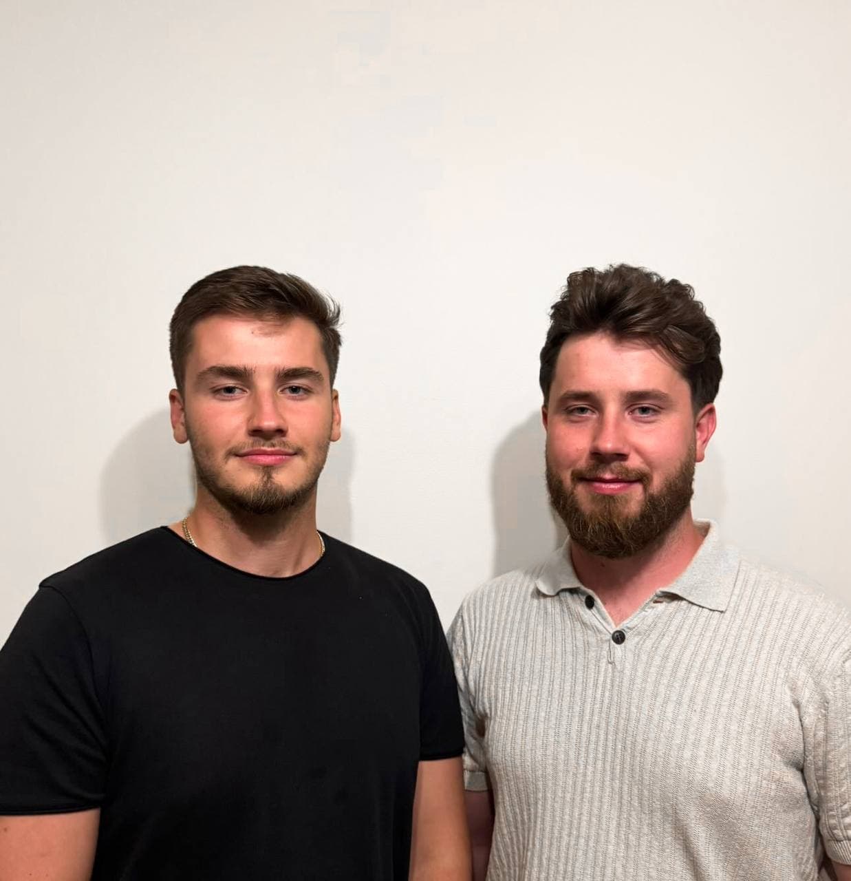 Two young men standing side by side and smiling in front of a plain white wall. - Home Instead