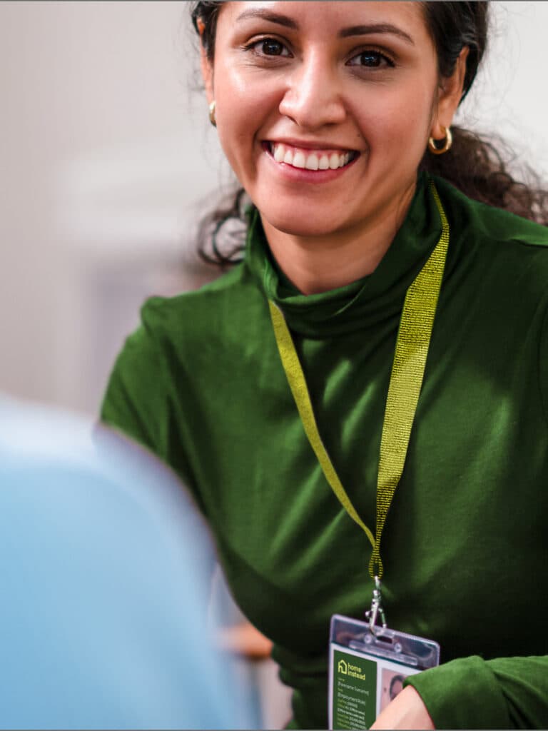 Woman in green top smiling, wearing a lanyard with an ID badge, sitting indoors. - Home Instead