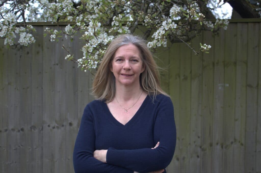 A woman with long blonde hair stands in front of a wooden fence and flowering tree, arms folded, smiling. - Home Instead